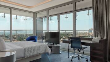 Quarto de hotel moderno com uma cama grande, uma escrivaninha, uma cadeira, uma televisão e vista para a cidade.