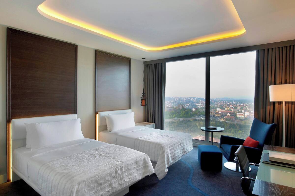 A hotel room with two beds, a view of a cityscape, a modern chair, and a small table.