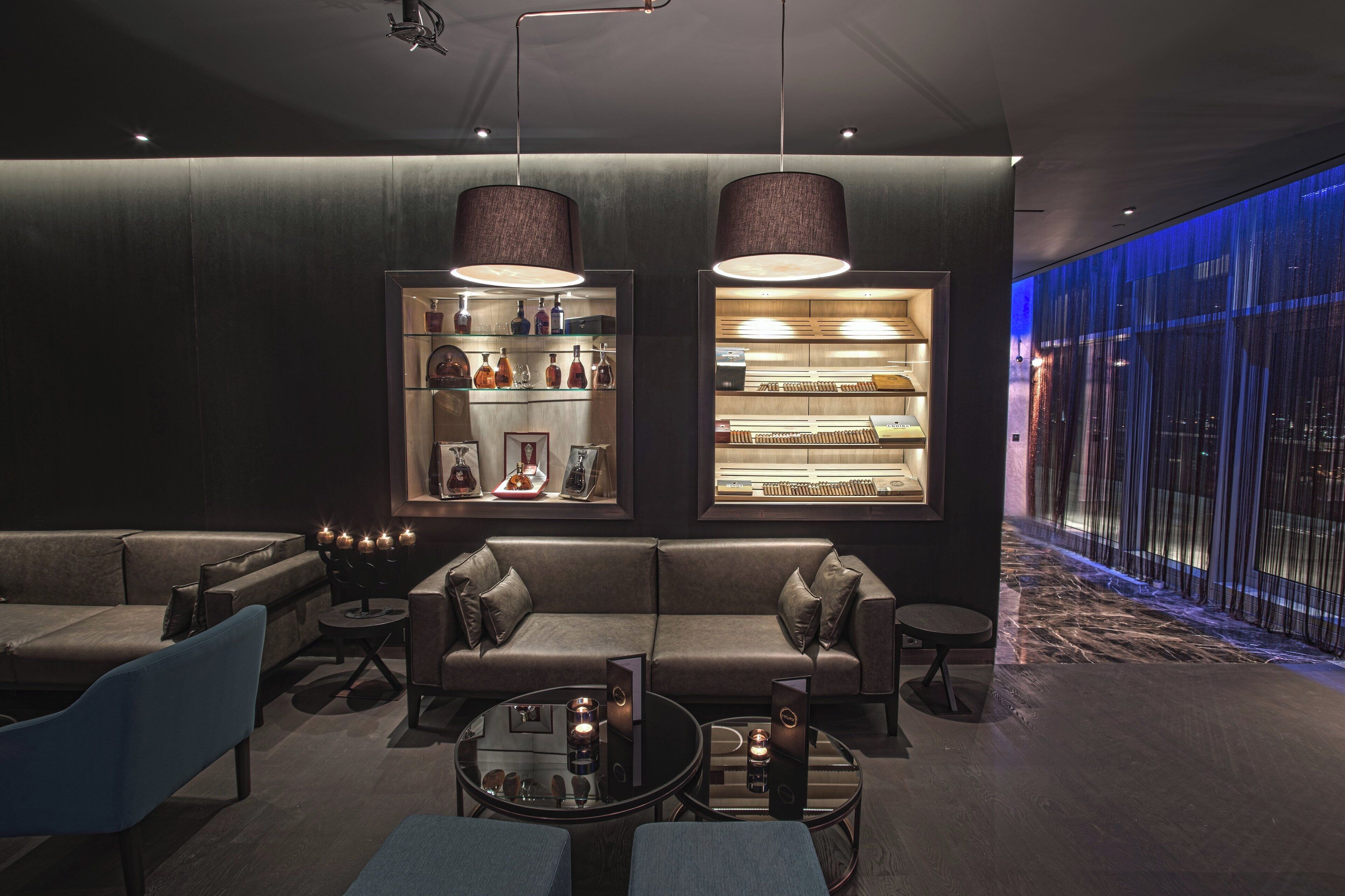 Ein moderner Lounge-Bereich mit einer Couch, Sesseln und einem Couchtisch. Zwei Pendelleuchten und eine Vitrine mit Gegenständen sind vorhanden.