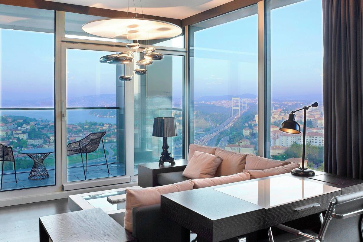 A modern living room with a sofa, a coffee table, and a dining table. There is a large window with a view of a cityscape and a bridge.