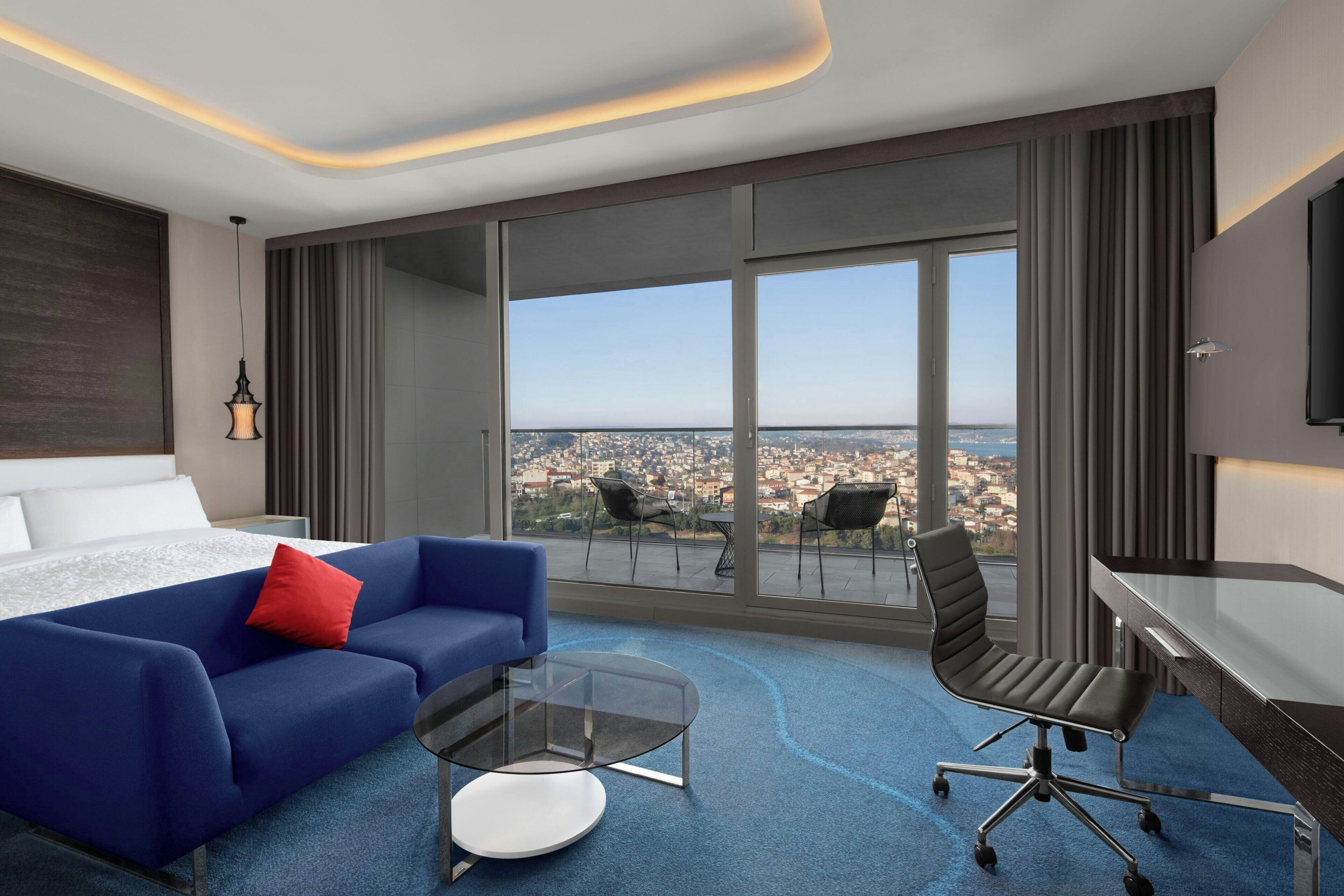 Una camera d'albergo moderna con un letto grande, un divano blu, un tavolino in vetro, una scrivania e una sedia. La stanza dispone di una grande finestra con vista sulla città.