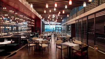 Un restaurant moderne avec plusieurs espaces de restauration, des luminaires suspendus et une vue sur le paysage urbain grâce à de grandes fenêtres.