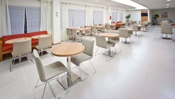 A modern dining area with round tables and chairs, a red sofa, and large windows with curtains.
