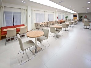 Un'area pranzo moderna con tavoli e sedie rotonde, un divano rosso e ampie finestre con tende.