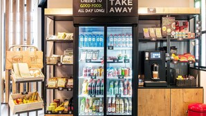 Un refrigerador para bebidas con un cartel arriba, exhibición de aperitivos y bebidas, y una estación de café con un cubo de basura rojo.