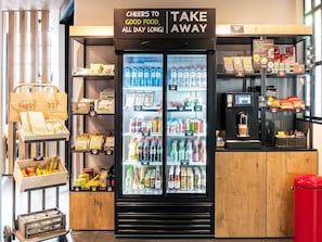 Een drankkoelkast met een bord erboven, een presentatie van snacks en drankjes, en een koffiehoek met een rode prullenbak.