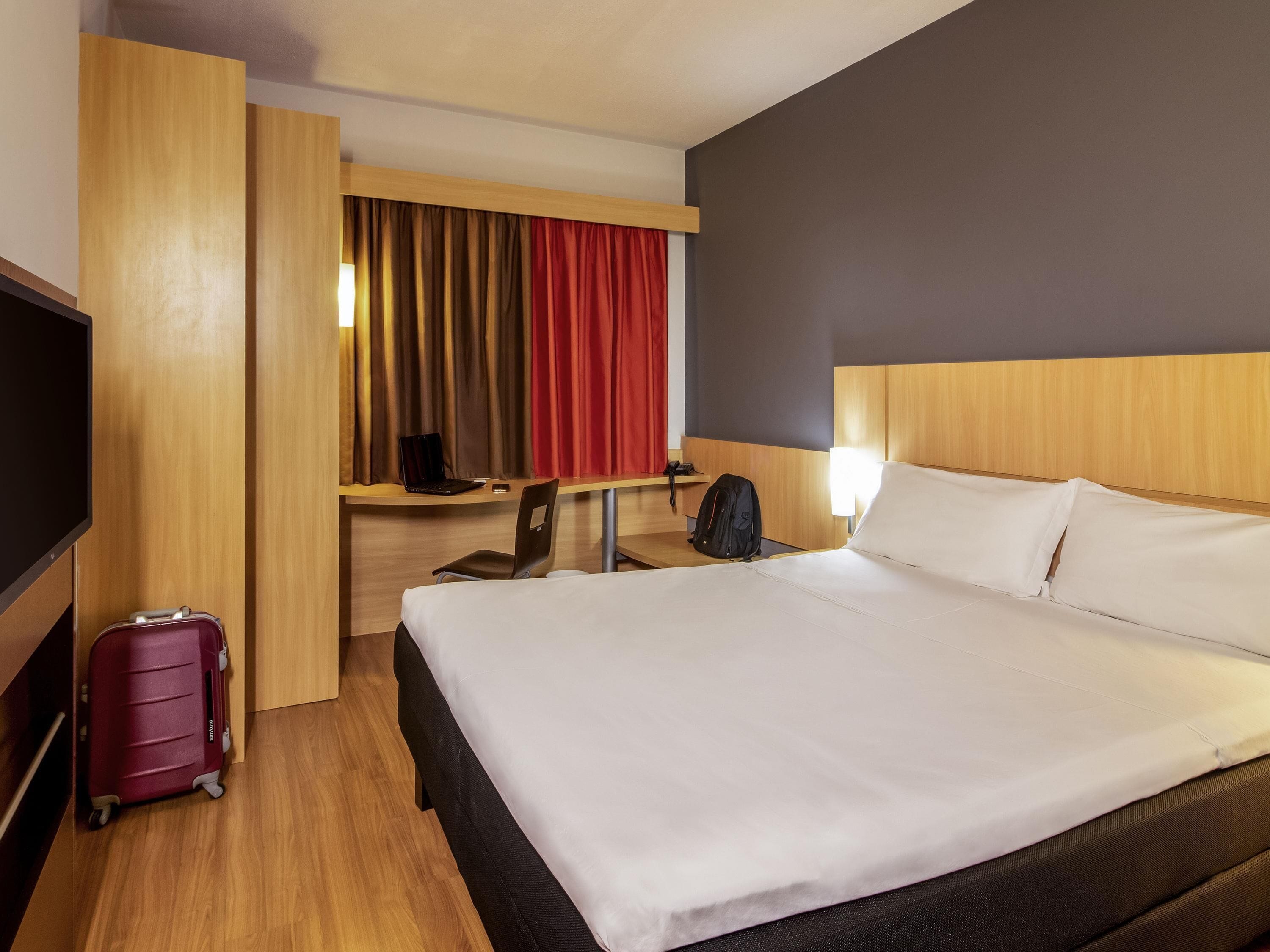 Une chambre d’hôtel avec un lit, un bureau avec un ordinateur portable, une chaise, une valise et un téléviseur.
