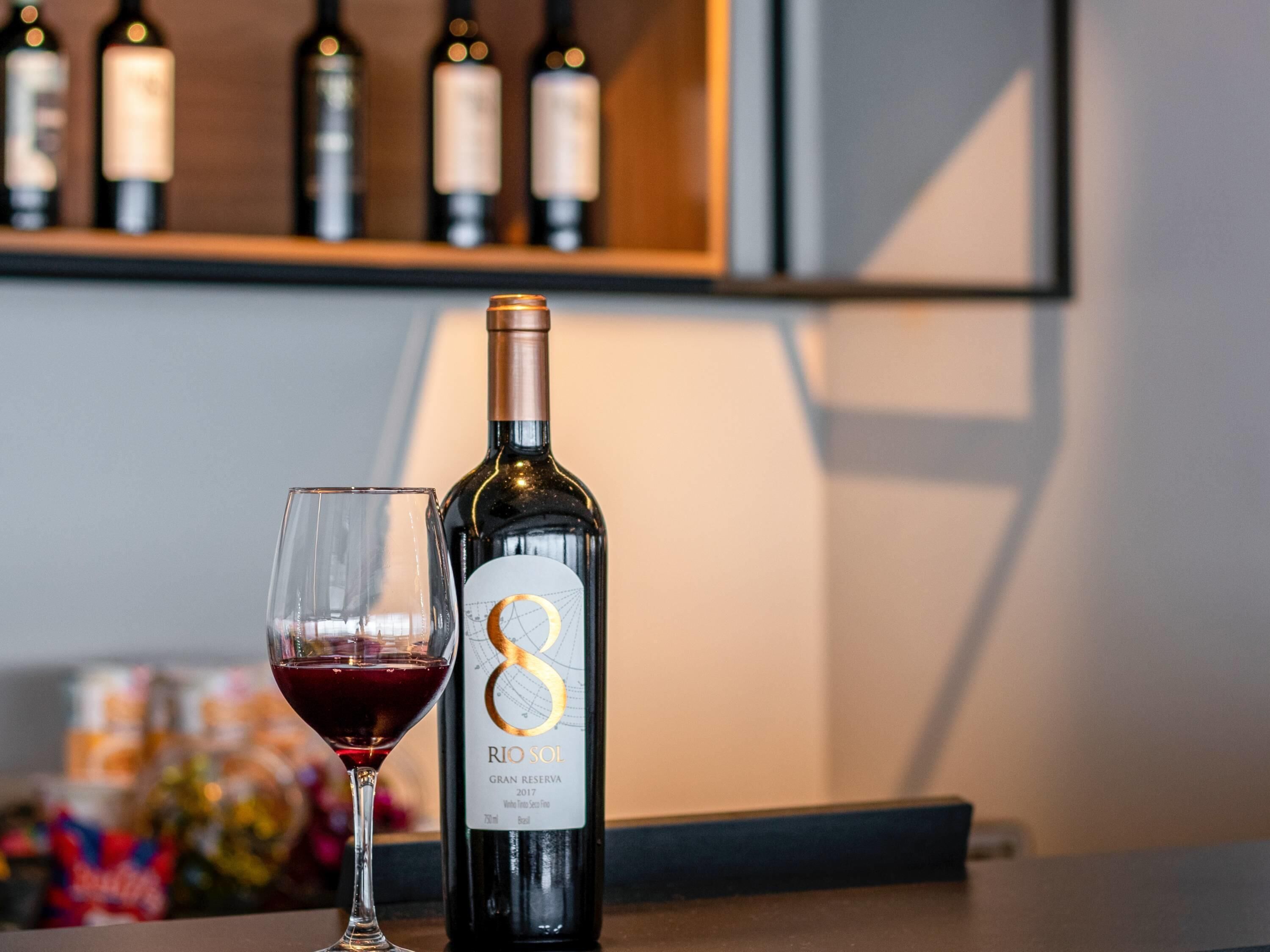 En flaska Rio Sol Gran Reserva 2017-vin står bredvid ett glas fyllt med rött vin, och i bakgrunden syns en hylla med vinflaskor.