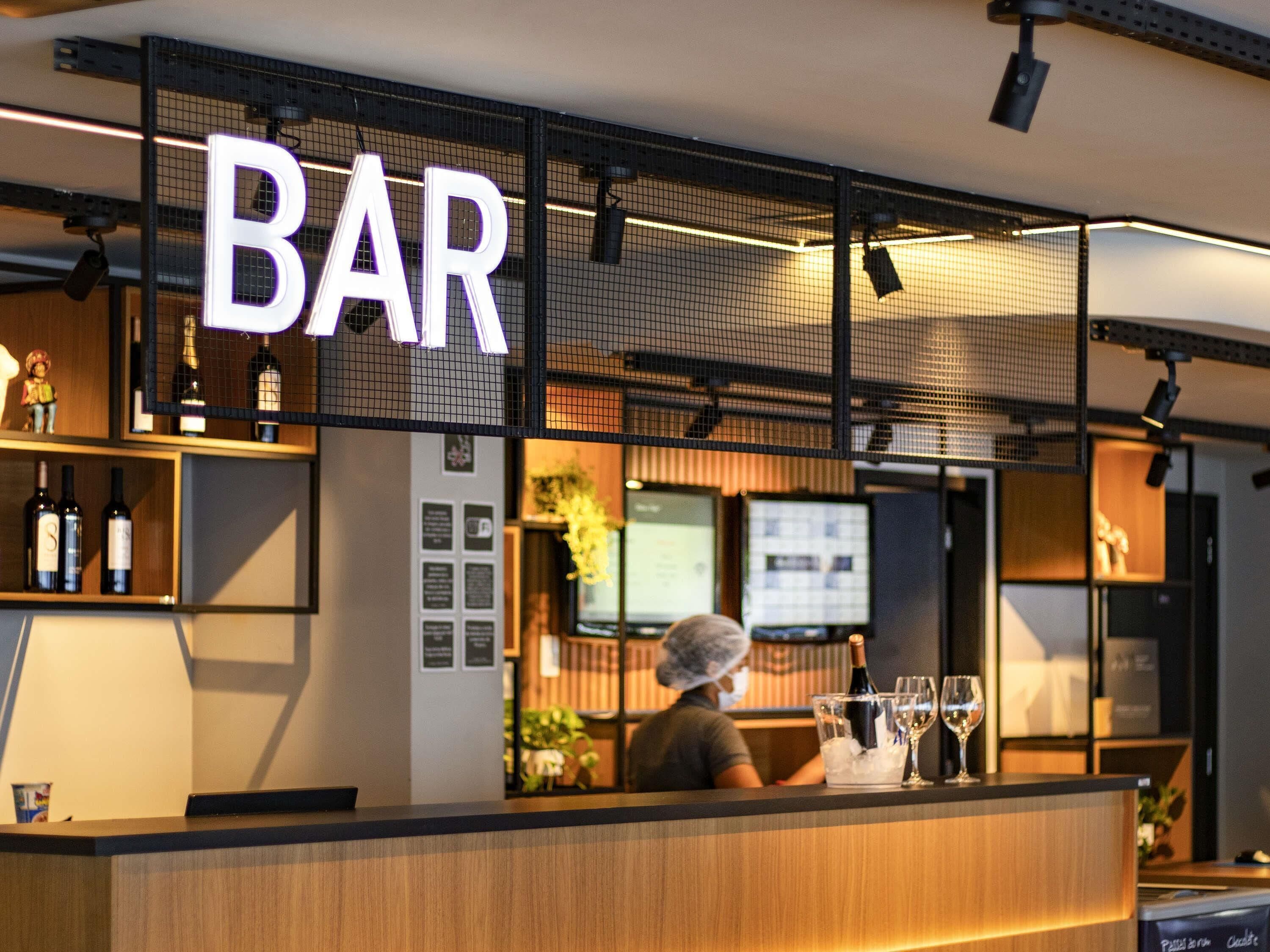 Zona de bar con un letrero que dice 'BAR', estantes con botellas, una barra y una persona sirviendo.