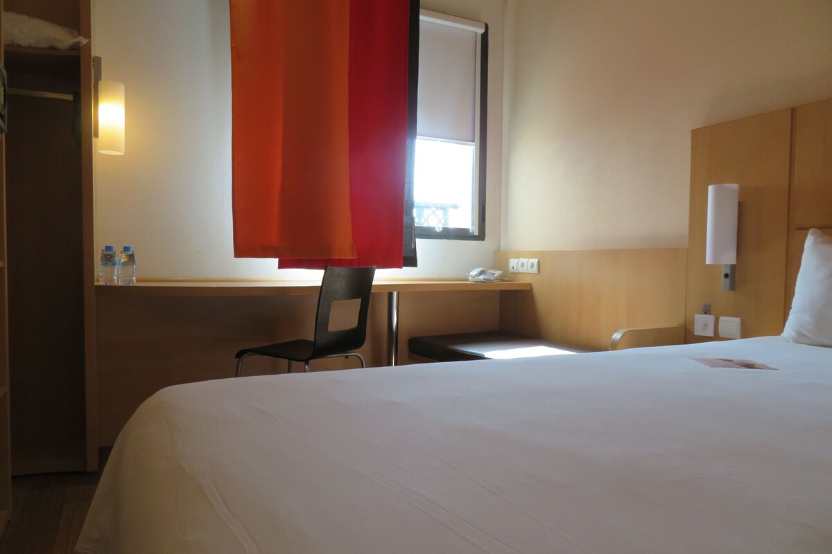 A hotel room with a large bed, a desk with a chair, a window with a red curtain, and a bedside table with a phone and a lamp.