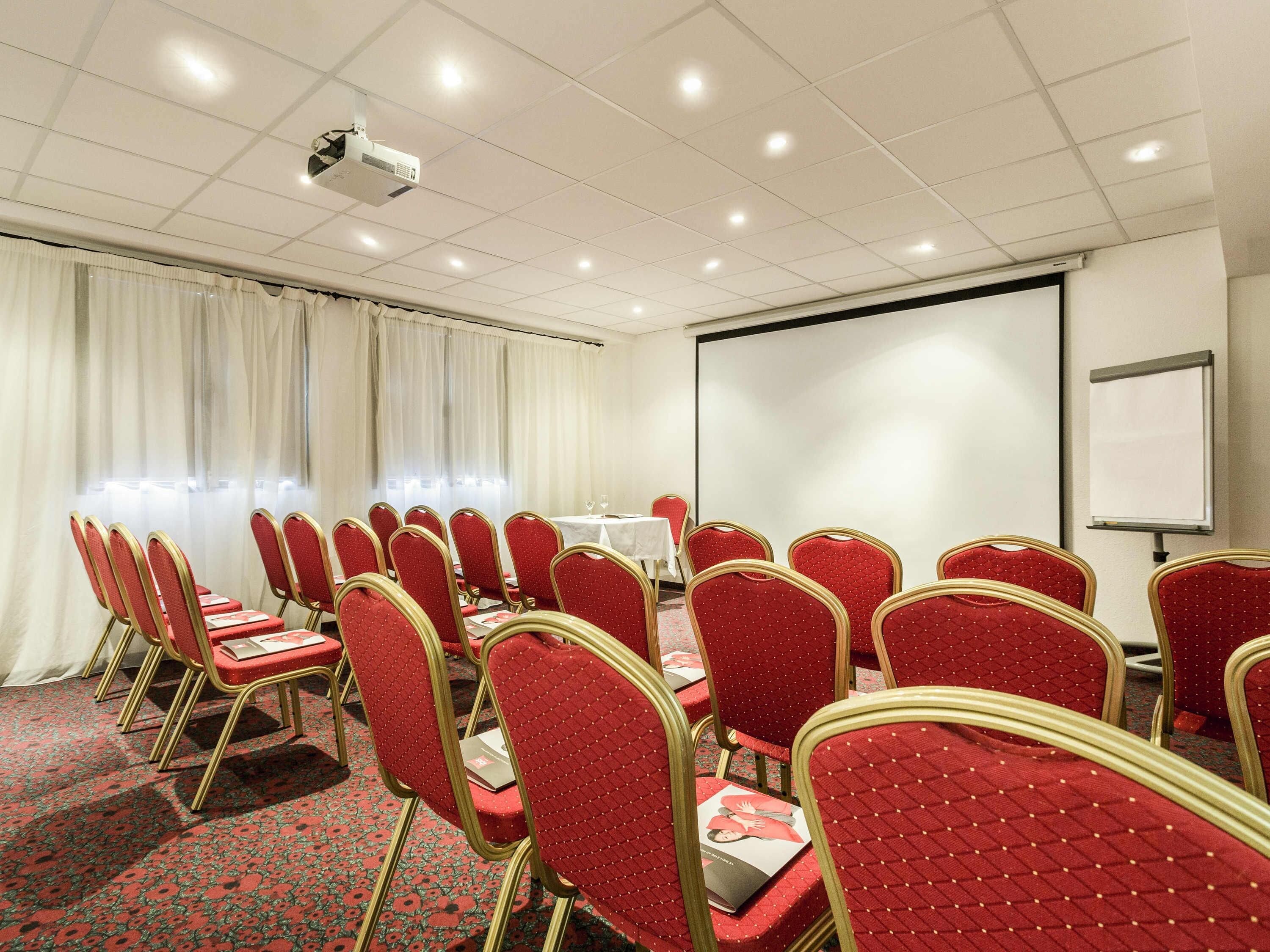 Un salón de conferencias con filas de sillas rojas, un proyector, una pantalla blanca y una alfombra con motivos florales.