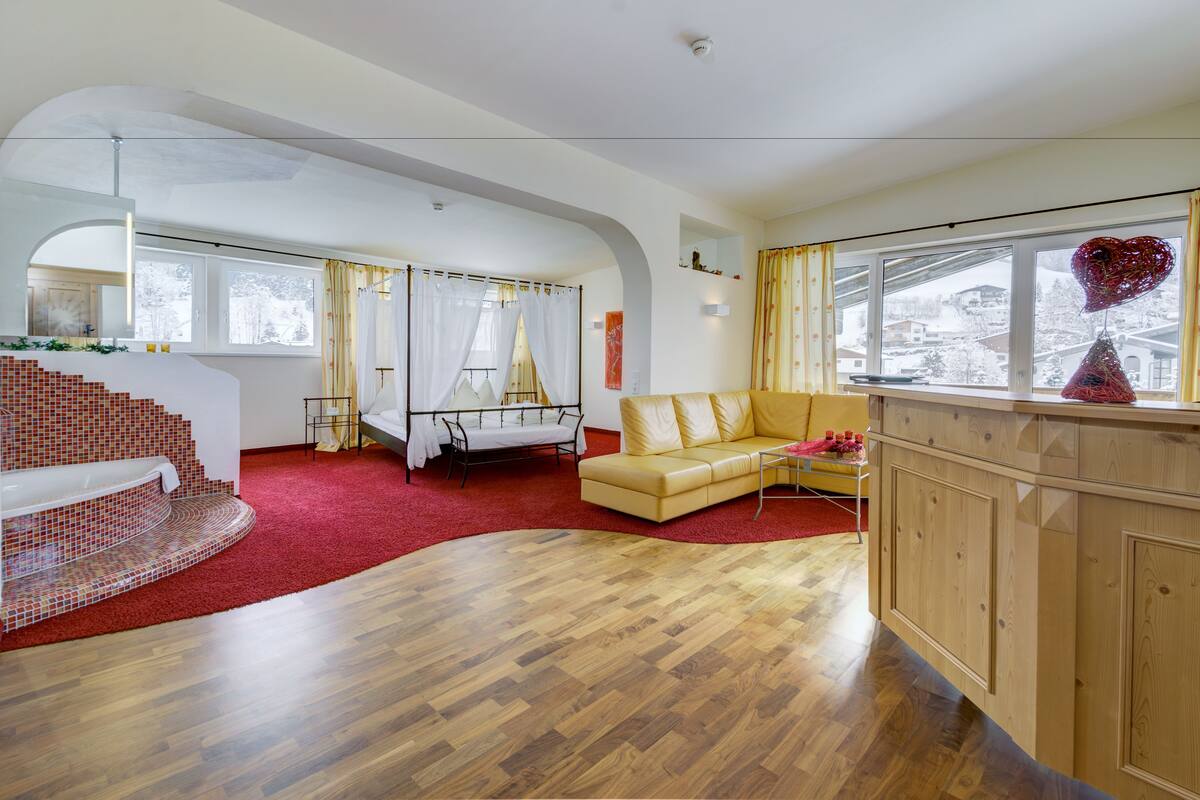 A spacious living room with a wooden floor, a yellow sofa, a white armchair, a red carpet, a staircase with red tiles, and a large window with a heart-shaped decoration.