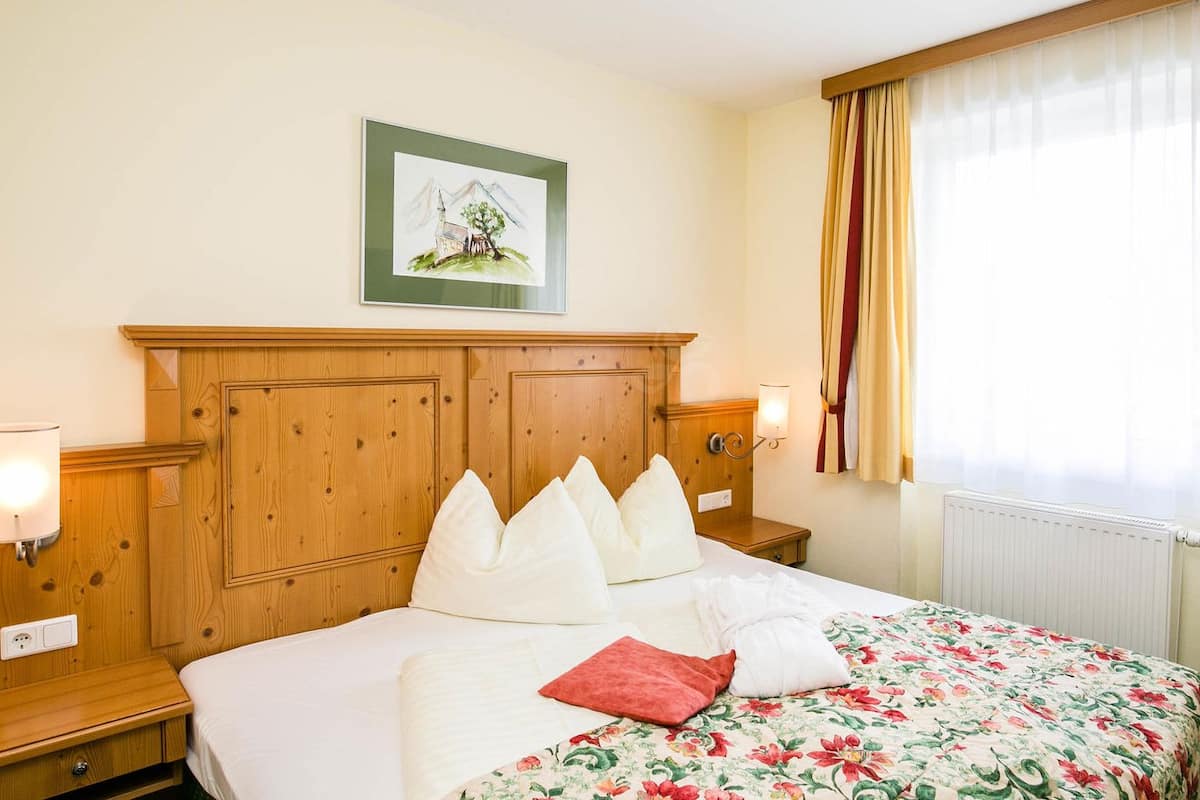 A hotel room with a wooden headboard, a bed with white bedding and a floral-patterned duvet, a nightstand with a lamp, and a framed picture above the bed.