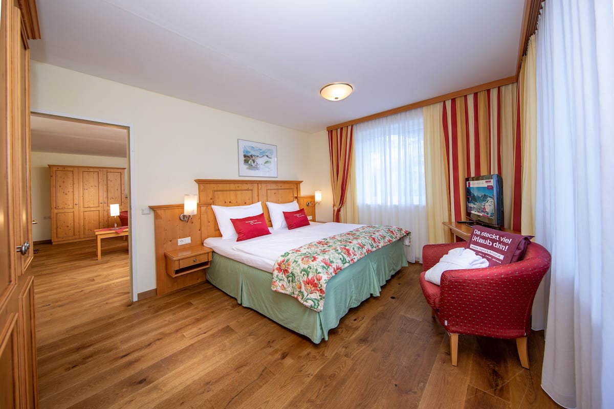 A hotel room with a wooden floor, a bed with green bedding, a red armchair, and a wooden wardrobe.