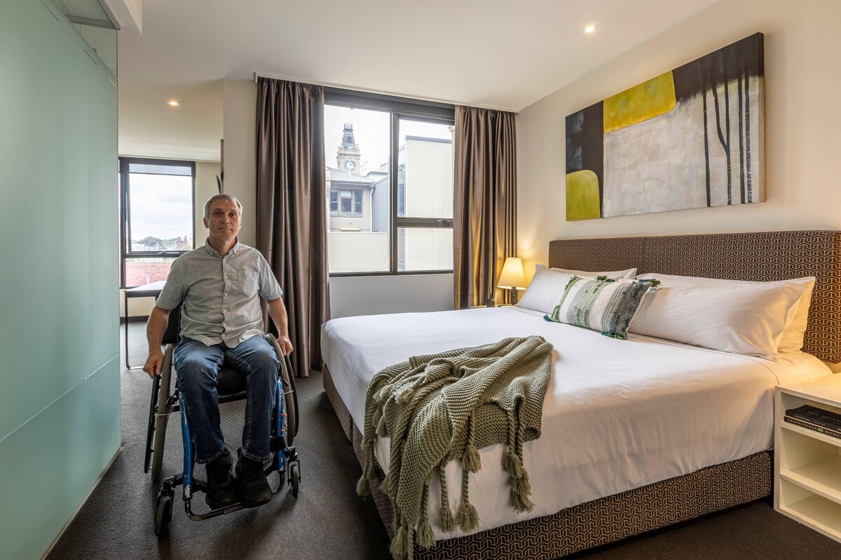 A hotel room with a bed, a wheelchair, a window with curtains, and a painting on the wall.