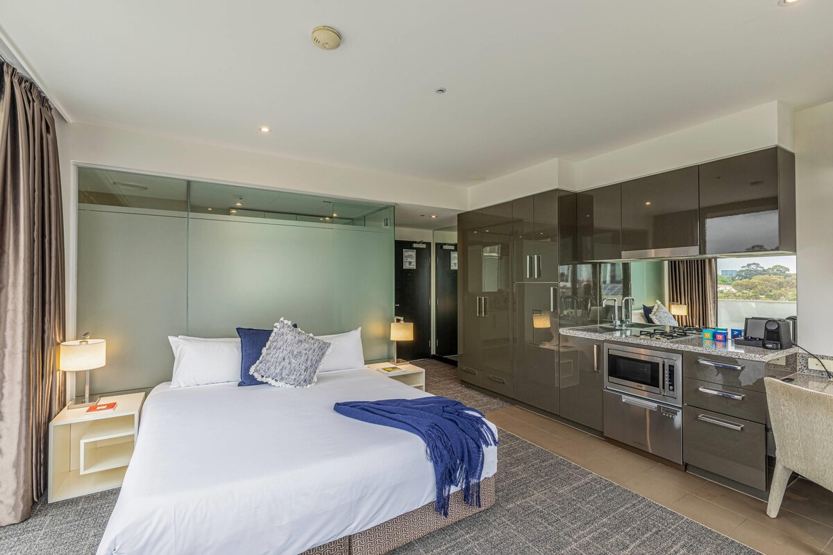 A modern hotel room with a large bed, a separate kitchenette, and a view through a glass door.