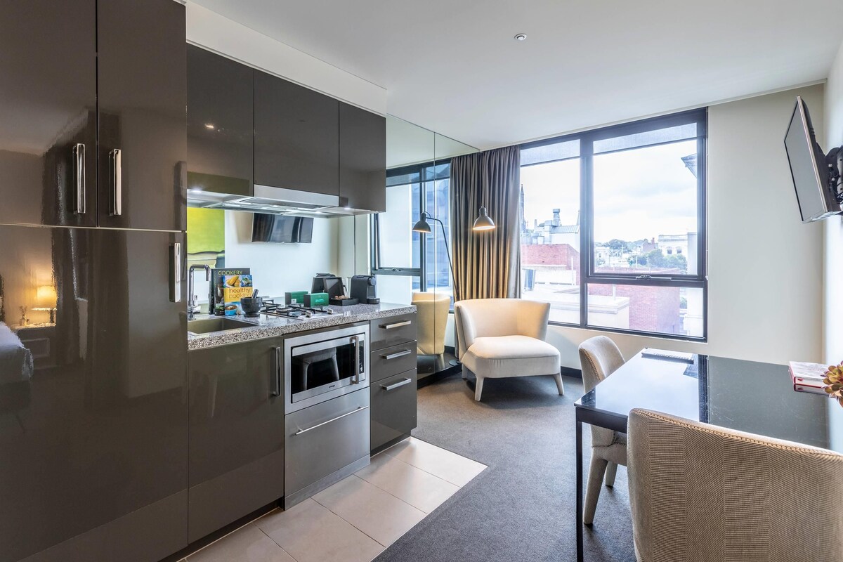 A modern kitchen with stainless steel appliances, a dining area with a table and chairs, and large windows offering a city view.