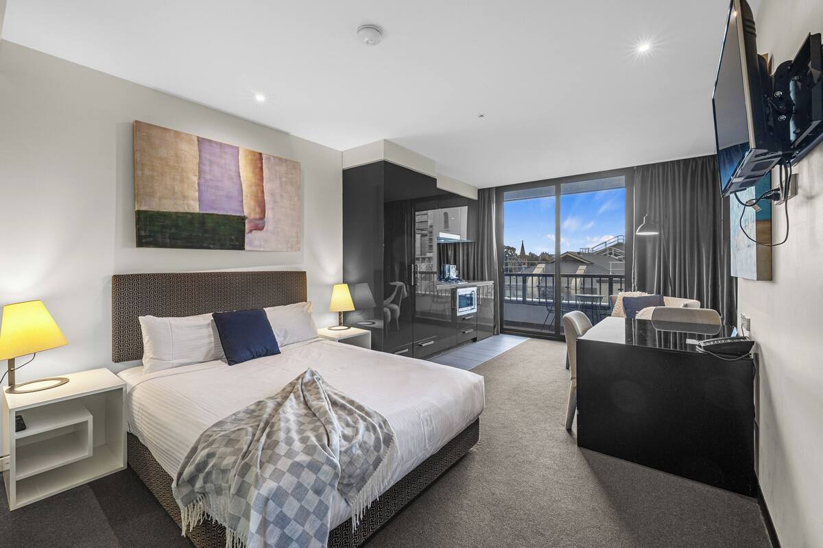 Quarto de hotel moderno com uma cama grande, uma escrivaninha, televisão e varanda com vista para a cidade.