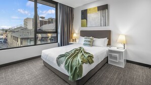 Ein Hotelzimmer mit großem Fenster, einem Bett mit grüner Tagesdecke, zwei Nachttischen mit Lampen und einem modernen Wandbild.