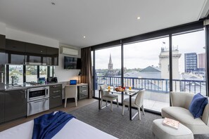 Habitación de hotel moderna con kitchenette, comedor con desayuno, ventana grande con vista a la ciudad y balcón con barandilla.
