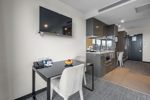 A modern hotel room with a dining area, a flat-screen TV, a small table with a bowl of fruit, and a kitchenette.