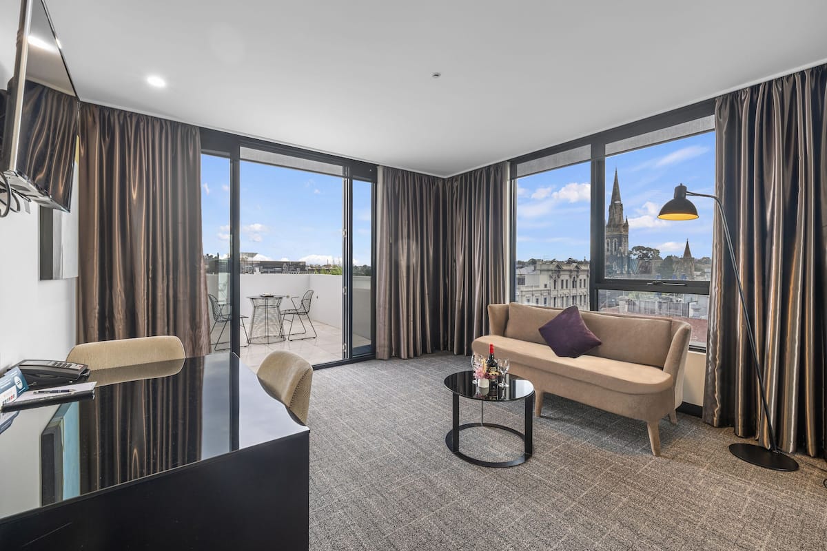 Quarto de hotel moderno com sofá, mesa de centro, escrivaninha e janelas amplas com vista para a cidade.