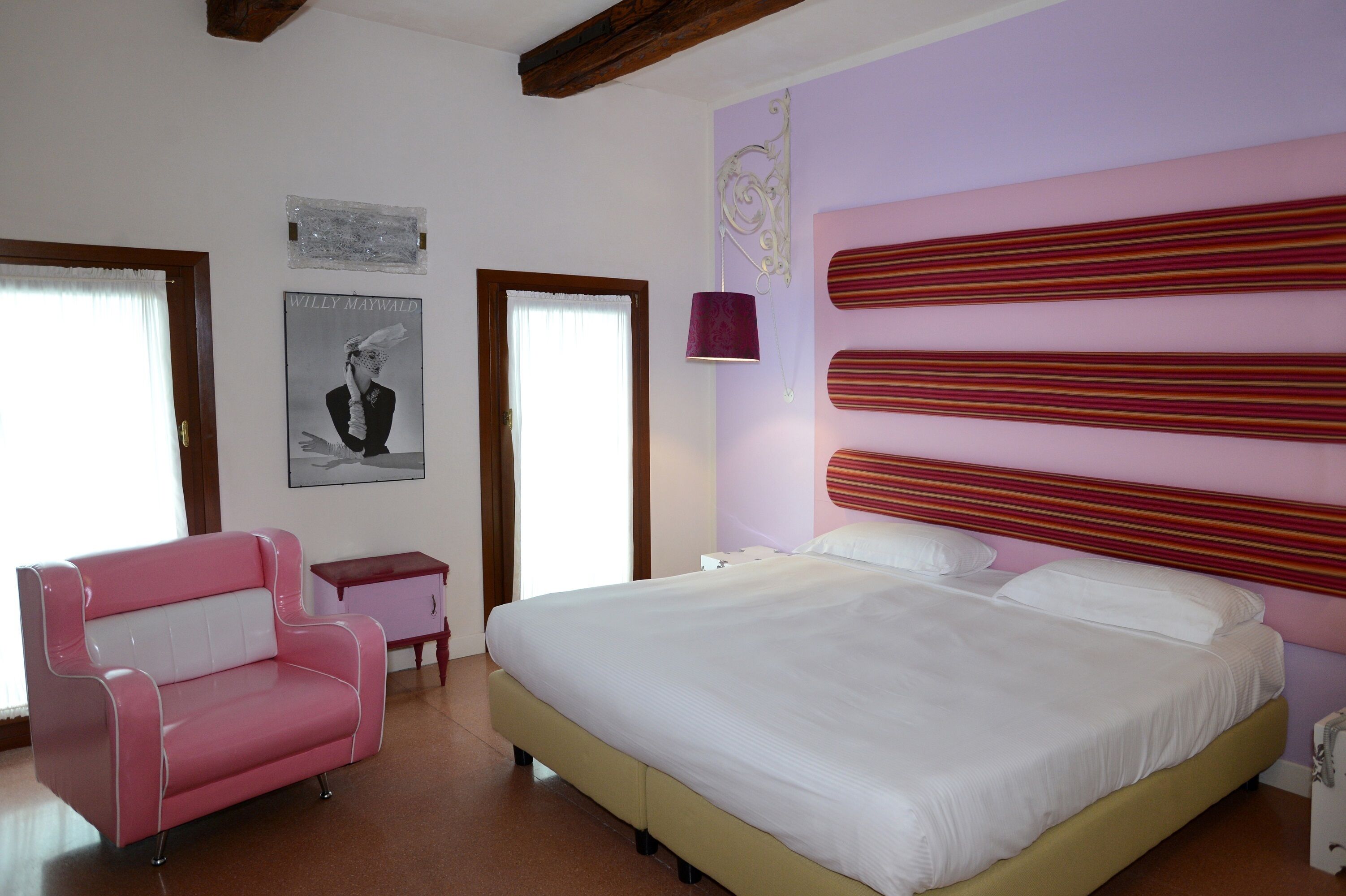 A bedroom with a large bed, a pink chair, a small desk, and a framed picture on the wall.
