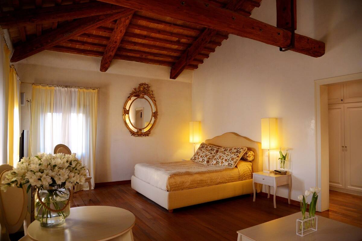 Ein Schlafzimmer mit Bett, Nachttischlampen, einem Spiegel, einer Vase mit Blumen und Holzbalken an der Decke.