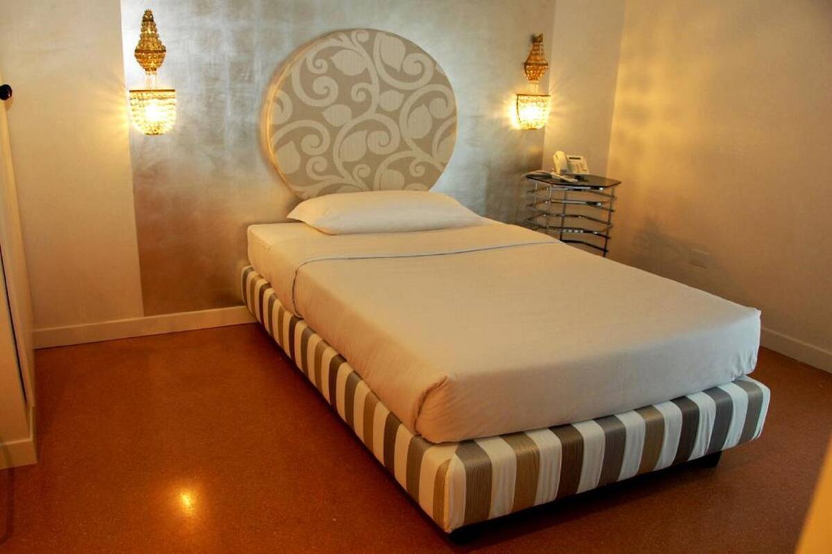 Ein ordentlich bezogenes Bett mit gestreifter Fußleiste und Kopfteil, ein Nachttisch mit Lampe sowie ein an der Wand befestigtes Telefon.
