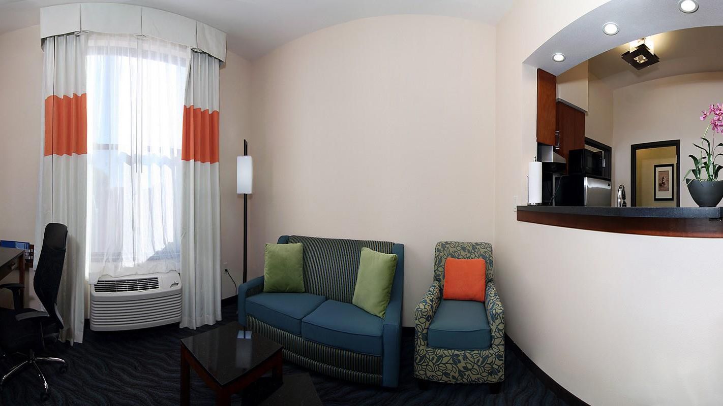 A hotel room with a sofa, armchair, and a small table. There is a kitchen area with a microwave and a sink.