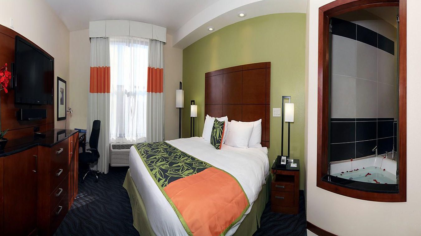 Een hotelkamer met een groot bed, een bureau met stoel, een televisie, een raam met gordijnen, een nachtkastje met lamp en een badkamer met bad.