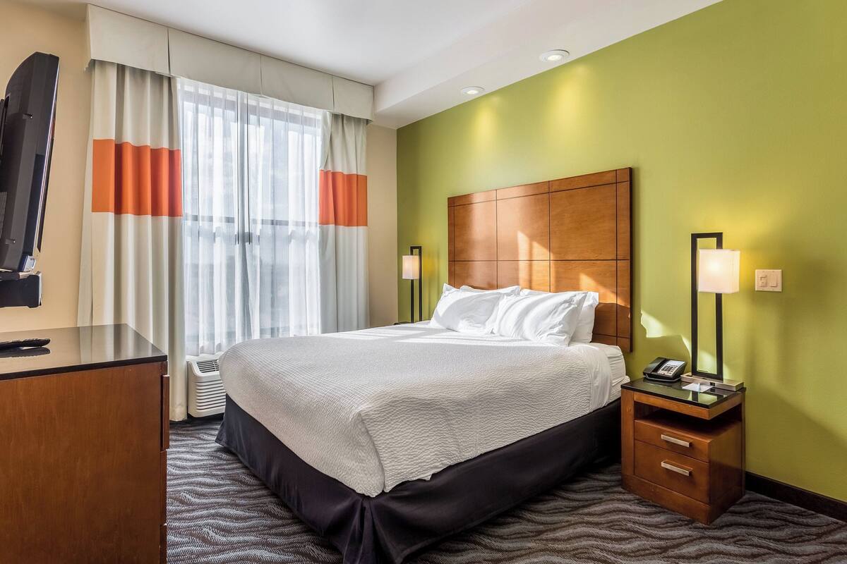 A hotel room with a bed, a desk, a chair, a TV, and a nightstand with a lamp.