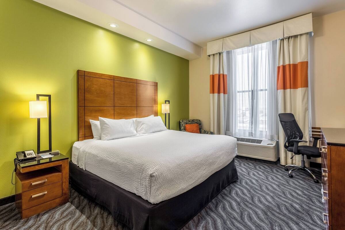 A hotel room with a large bed, a desk with a chair, a nightstand with a lamp, and a window with curtains.