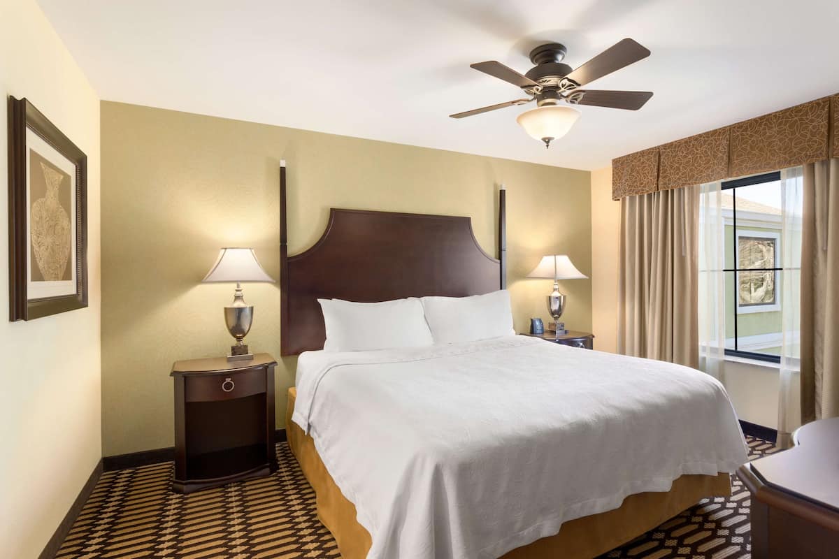 Habitación de hotel con una cama grande, dos mesitas de noche con lámparas, un ventilador de techo y una ventana con cortinas.