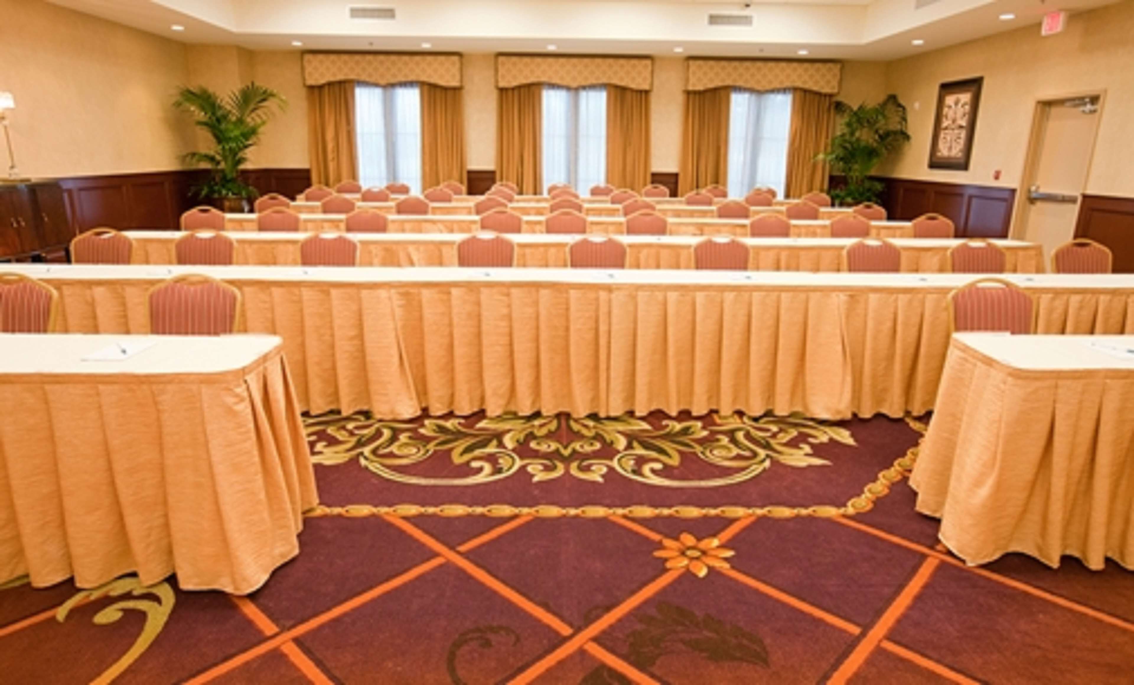 A conference room with rows of tables and chairs, a patterned carpet, and large windows with curtains.