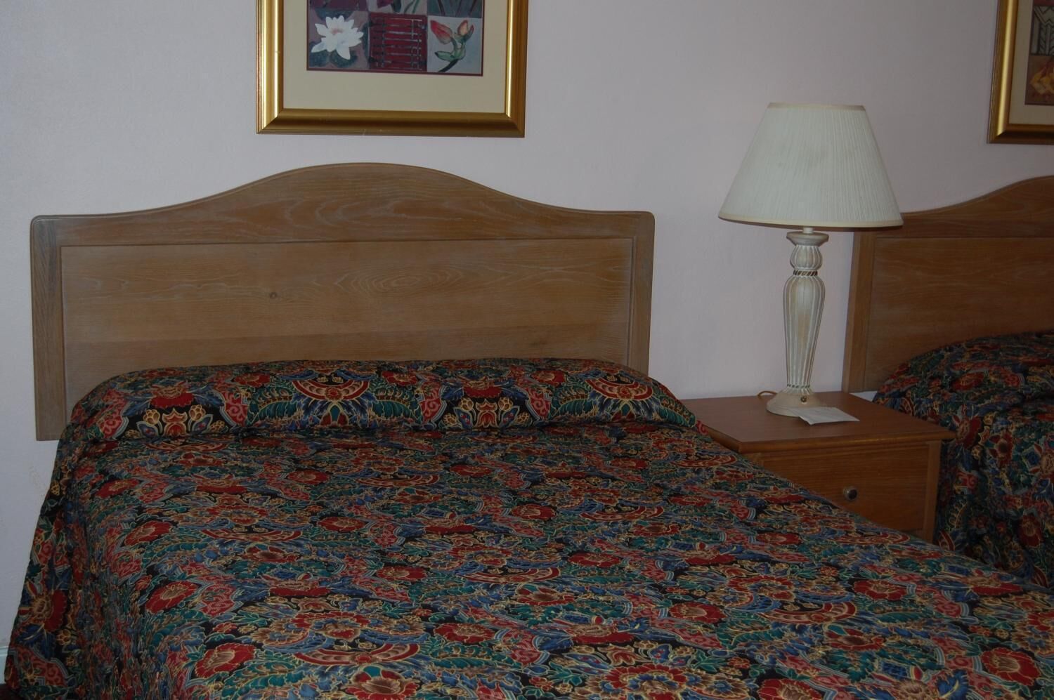 A room with a wooden headboard, a patterned bedspread, a bedside table with a lamp, and a framed picture on the wall.