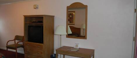 A hotel room with a wooden wardrobe, a television, a desk with a lamp, a chair, and a patterned bedspread.