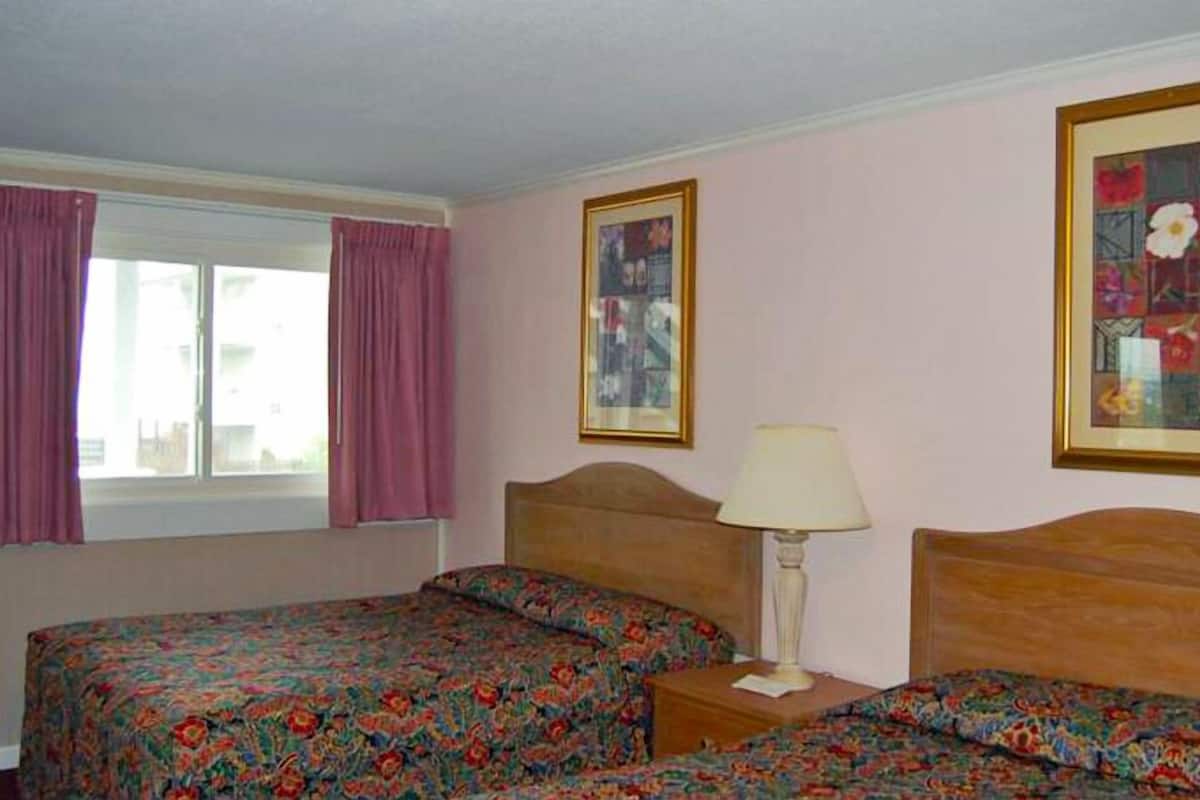 Una habitació d'hotel amb dos llits, una finestra amb cortines roses, un quadre al mur i una taula de nit amb una làmpada.