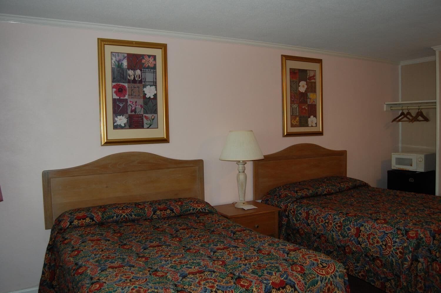 A room with two wooden beds, each with a patterned bedspread, a bedside table with a lamp, and a framed floral artwork on the wall.