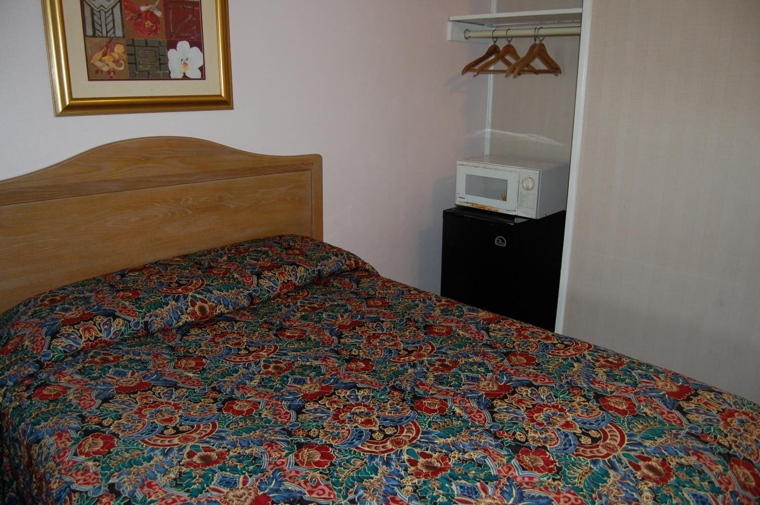 Une chambre avec une tête de lit en bois, une couvre-lit à motifs, un micro-ondes blanc posé sur un meuble noir et un tableau encadré accroché au mur.