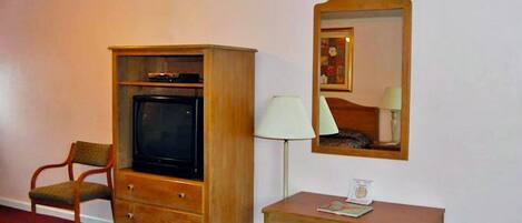 A room with a wooden TV cabinet, a chair, a small table with a lamp, and a mirror on the wall.