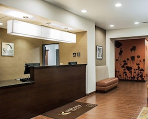A hotel reception area with a wooden reception desk, a sign for 'Sleep Inn & Suites', and a wall-mounted light fixture. - Sleep Inn & Suites Fort Campbell (Oak Grove)