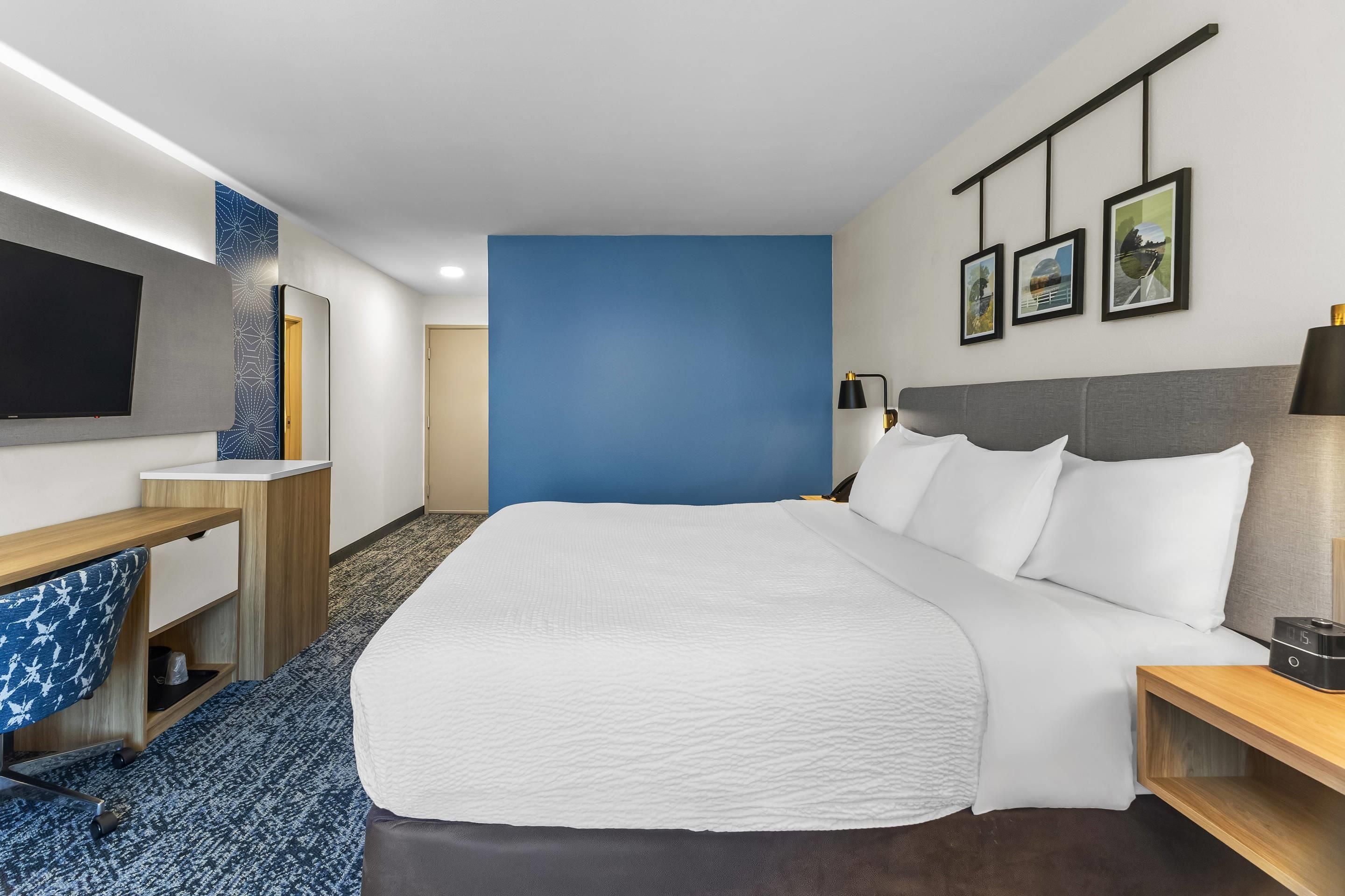 A hotel room with a large bed, a desk with a flat-screen TV, a wall-mounted lamp, and a framed picture above the bed.