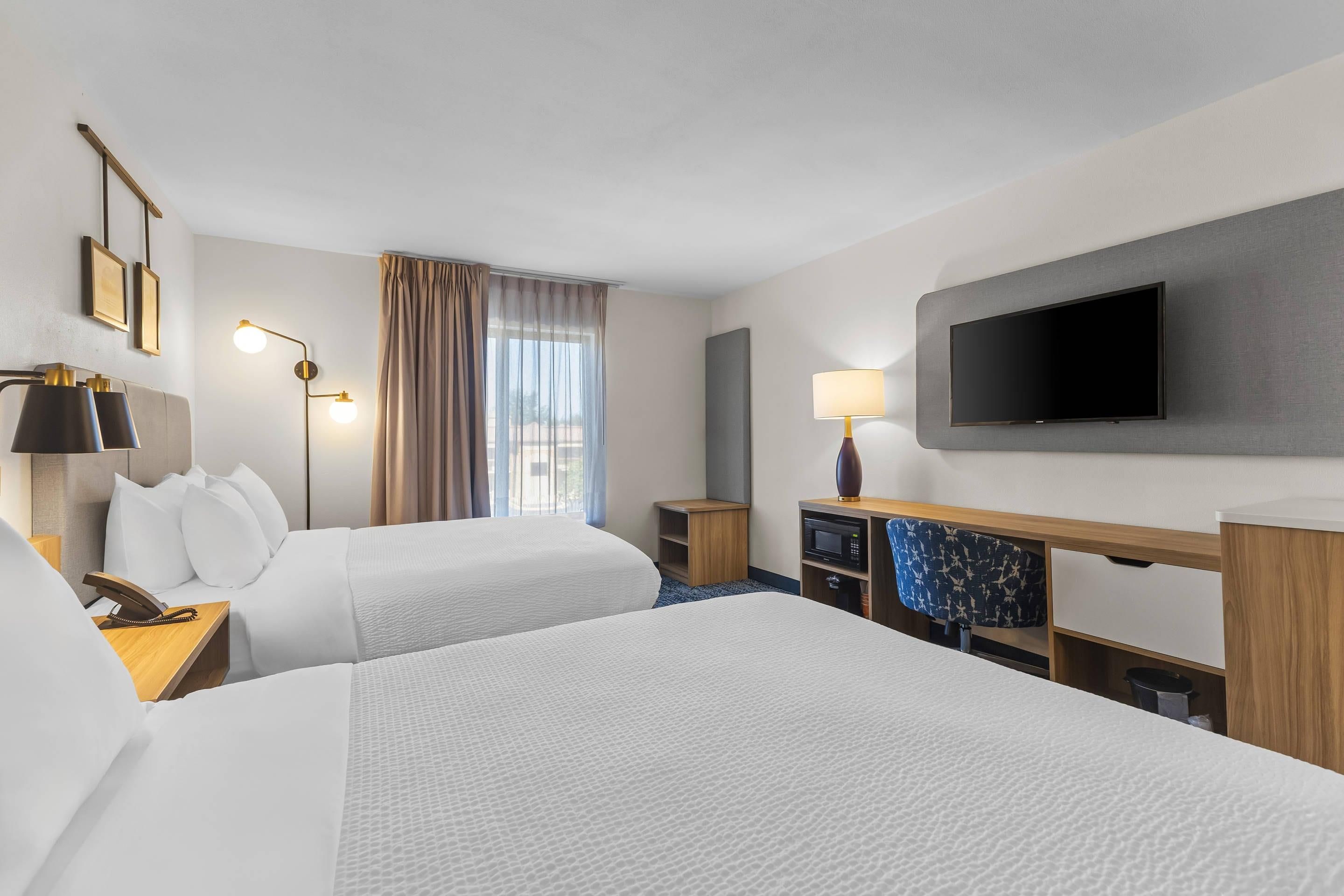 A hotel room with two beds, a desk with a telephone, a flat-screen TV mounted on the wall, and a window with curtains.