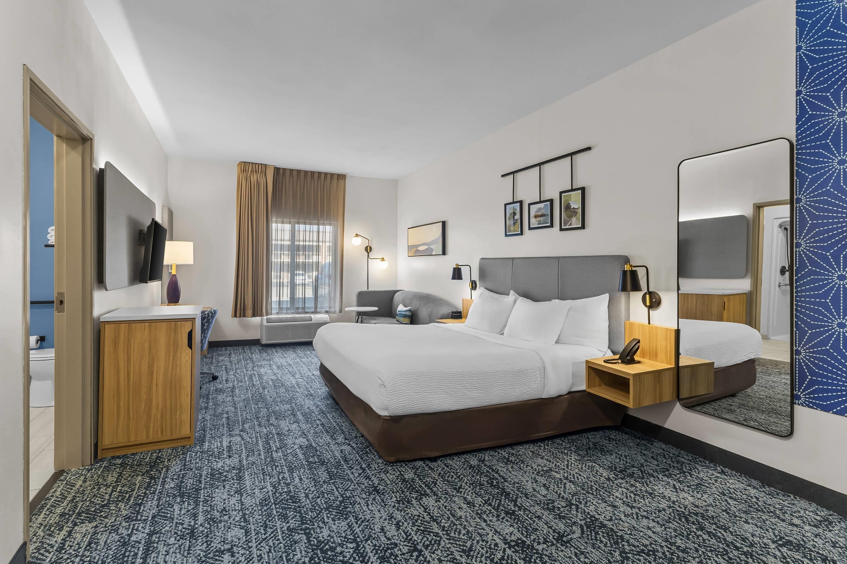 A hotel room with a bed, bedside tables, a desk, a chair, a TV, a window with curtains, and a mirror.