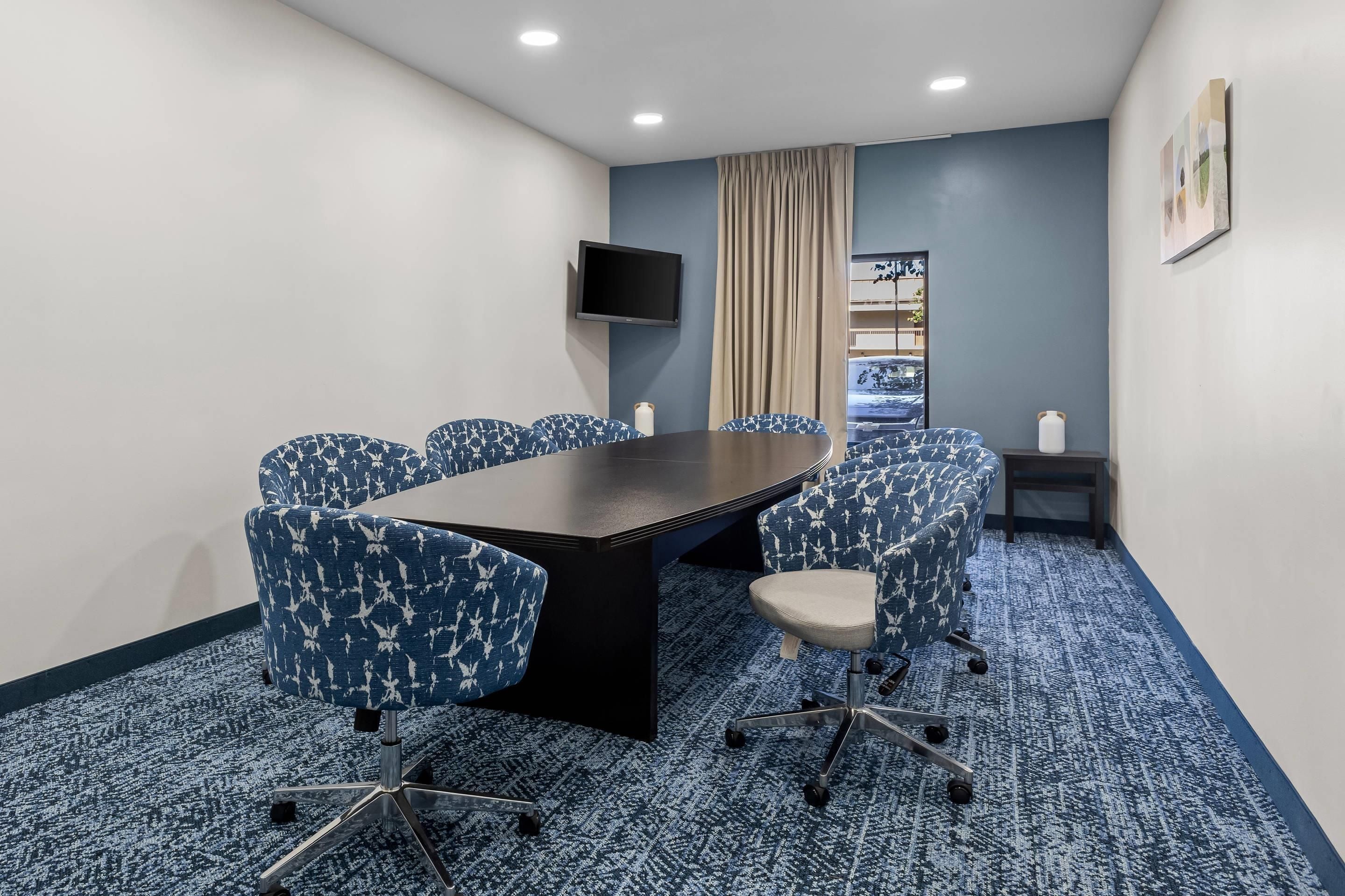A conference room with a long table, six chairs, a flat-screen TV, a window with curtains, and a view of a building.