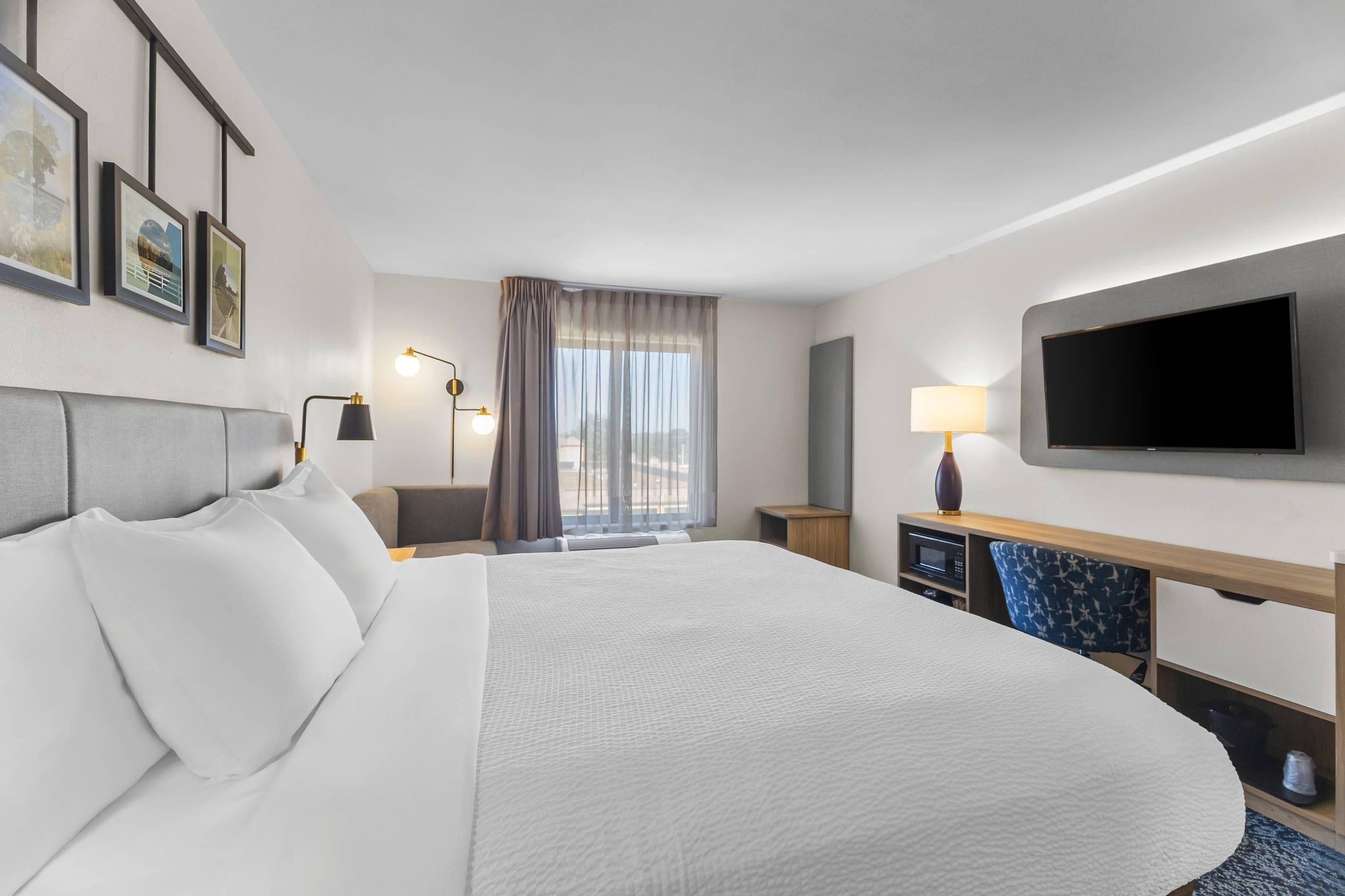 A hotel room with a large bed, a flat-screen TV, a desk with a lamp, and a window with curtains.