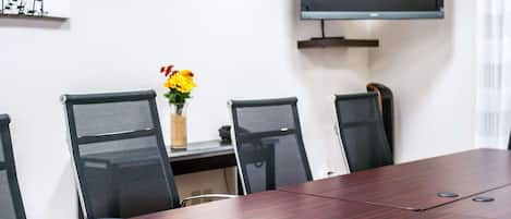 Un salón de conferencias con una larga mesa, cuatro sillas, un televisor en la pared y un florero con flores sobre una mesita.