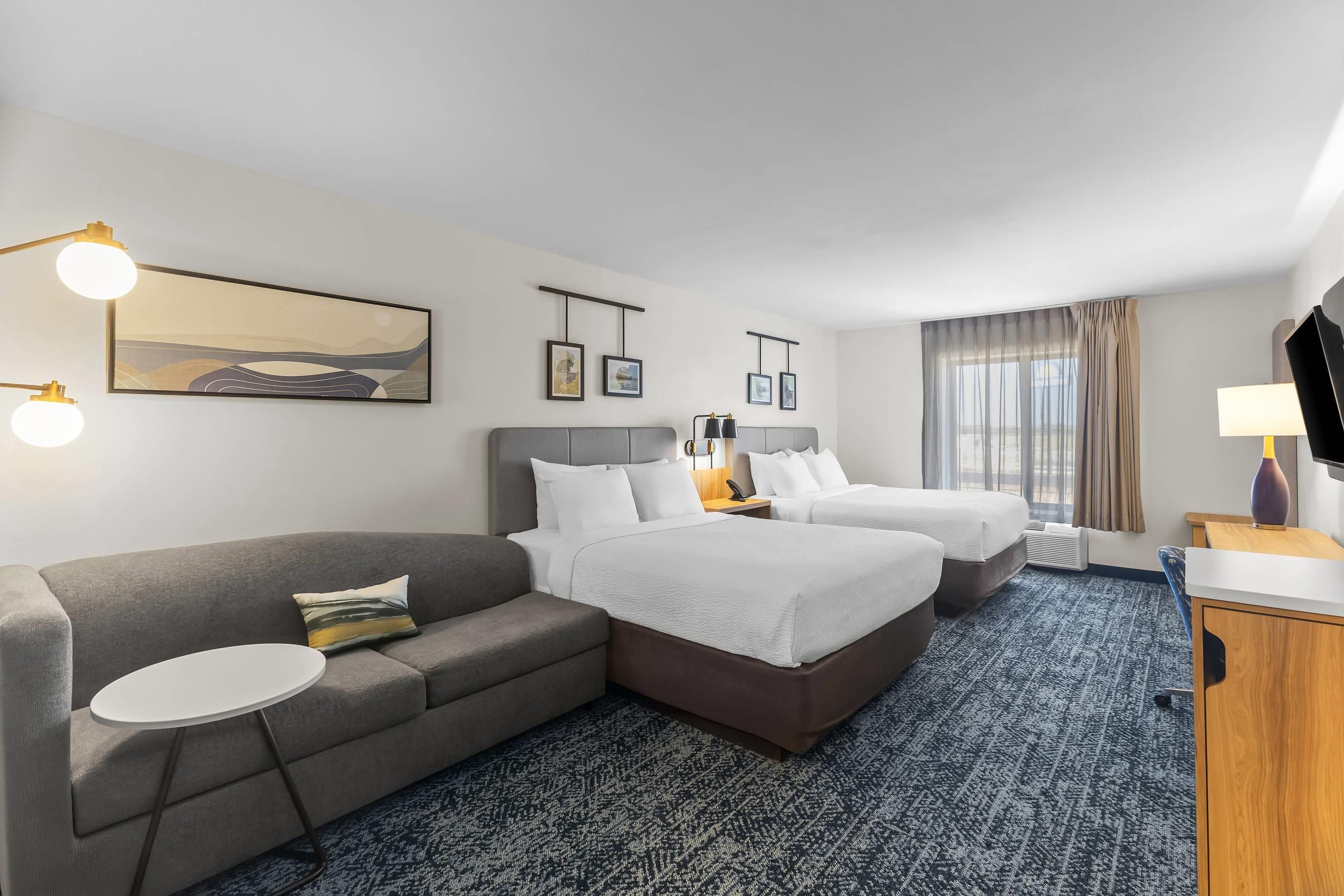 A hotel room with a grey sofa, two single beds, a small round table, and a TV on a wooden stand.