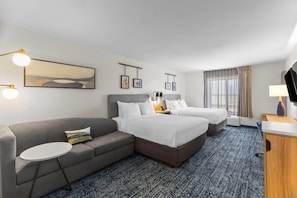 A hotel room with a grey sofa, two single beds, a small round table, and a TV on a wooden stand. - Sleep Inn & Suites Fort Campbell (Oak Grove)